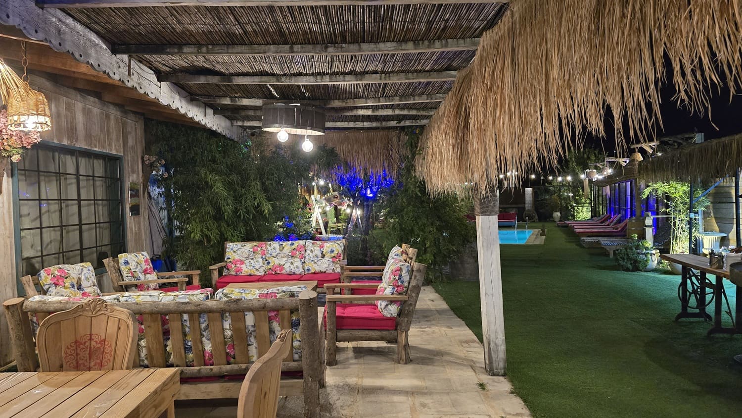 A cozy outdoor lounge and dining area at Kaya's Resort at night, featuring rustic log-style furniture, vibrant floral cushions, and a warm, inviting ambiance.