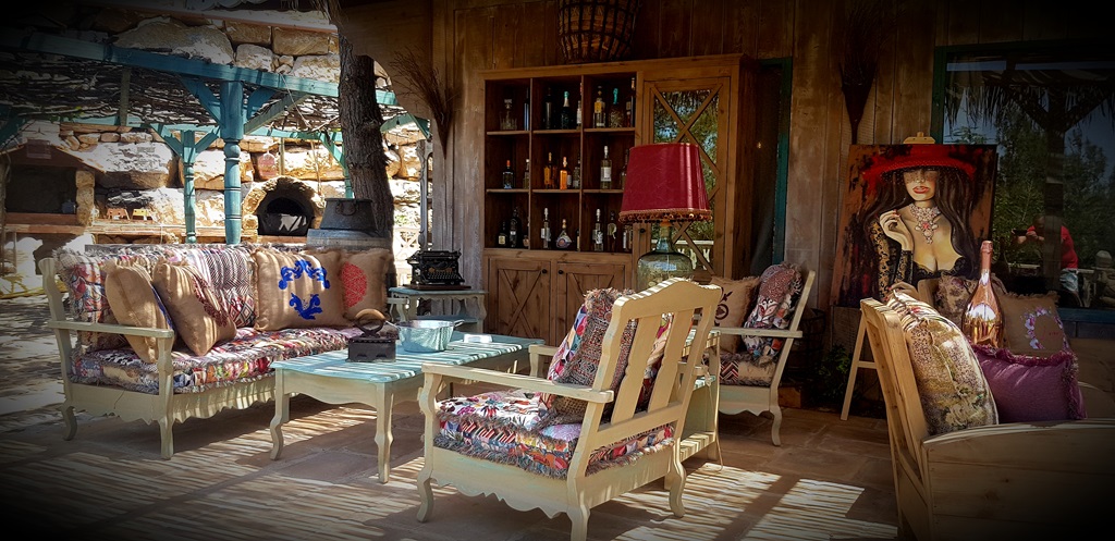 A rustic-chic outdoor lounge at Kaya's Resort featuring a wooden sofa with colorful bohemian cushions, a stone oven, and a vintage typewriter, creating a warm, artistic, and luxurious atmosphere.
