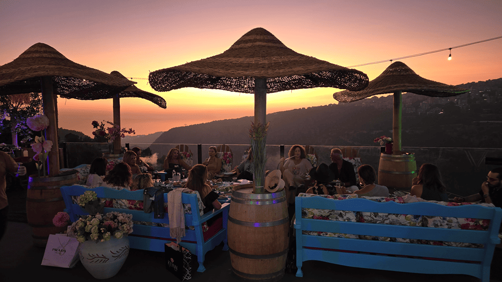 A group of friends enjoys the sunset from the vibrant rooftop lounge at Kaya's Resort, featuring blue benches, thatched umbrellas, and a scenic mountain view.